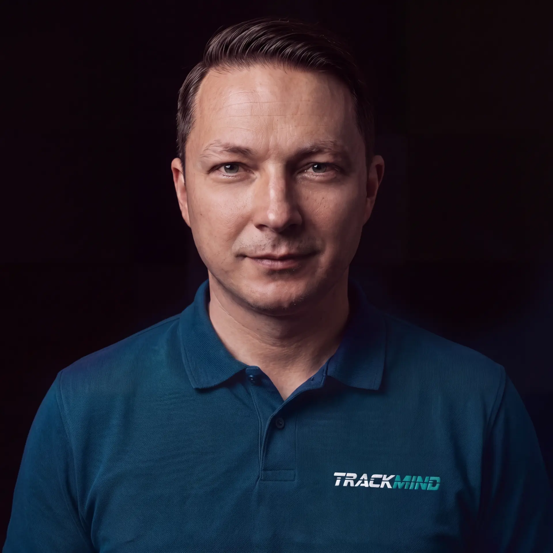 Porträt von Carlo Steckert, Gründer und Mental Performance Coach von TrackMind: kurze dunkle Haare, dunkelgrünes Polohemd mit TrackMind-Stickerei, vor dunklem Hintergrund, ruhiger Blick in die Kamera